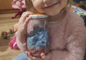 Uśmiechnięta dziewczynka trzyma wykonany w szklanym słoiku „kosmos” z przewagą niebieskiego koloru
